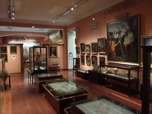 Fotografía donde se ve una de las salas del museo con vitrinas y pinturas expuestas.