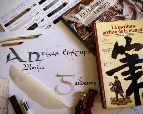 Imagen de una mesa de trabajo dedicada a la caligrafía: hay hojas con letras escritas a mano, plumas y herramientas de escritura antiguas, muestras de tinta y varios libros sobre escritura. Destaca un papel central con letras ornamentales y una pluma blanca, junto a sellos y materiales de práctica.